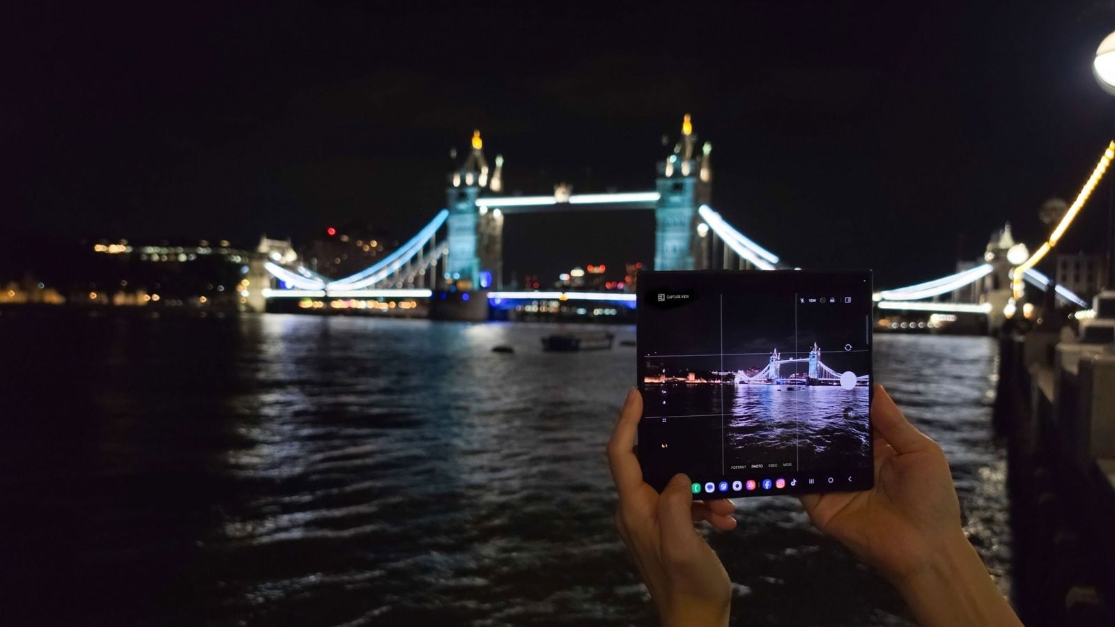 Ruky držia rozložený Samsung Galaxy Z Fold s otvorenou aplikáciou fotoaparátu, fotenie Tower Bridge v noci